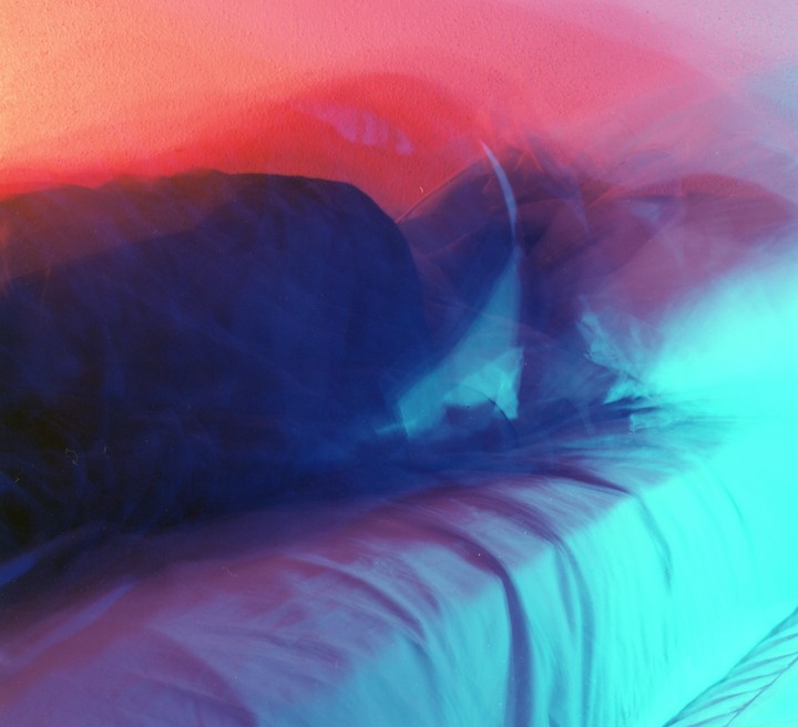A long exposure photograph shows a bed with a person sleeping under covers, illuminated by red and blue light.
