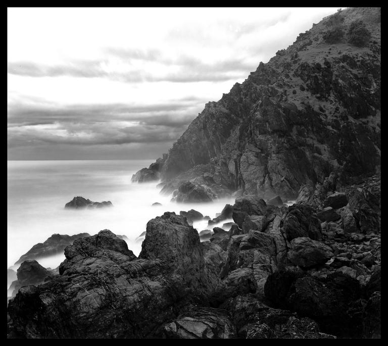 A rocky coastline with a large mountain on the right and blurred water on the left under a cloudy sky.