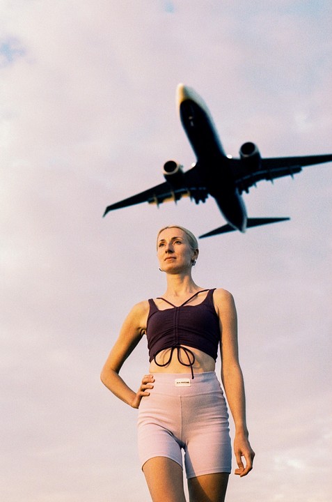 A woman stands with her hand on her hip as an airplane flies overhead.