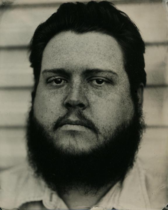 A man with a beard and freckles looks directly at the viewer in a close-up portrait.