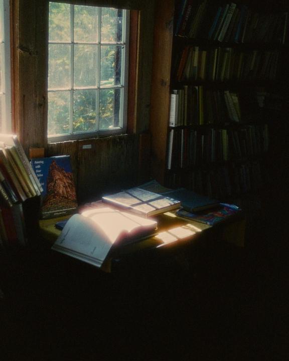 Sunlight streams through a window onto books on a table and a bookshelf in a dimly lit room.