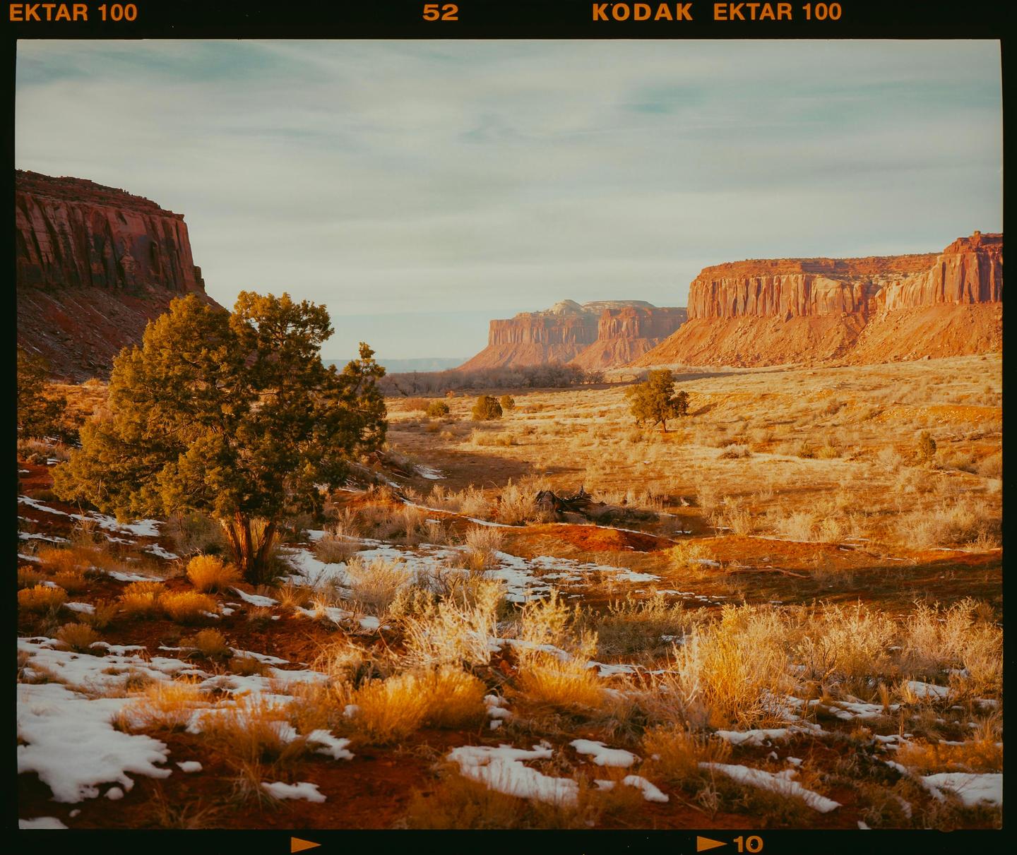 A desert landscape with red rock formations, sparse vegetation, and patches of snow is visible under a light sky.