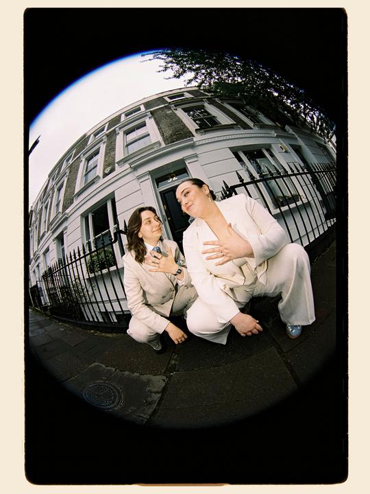 Two women in suits crouch on a sidewalk in front of a building with a black fence.