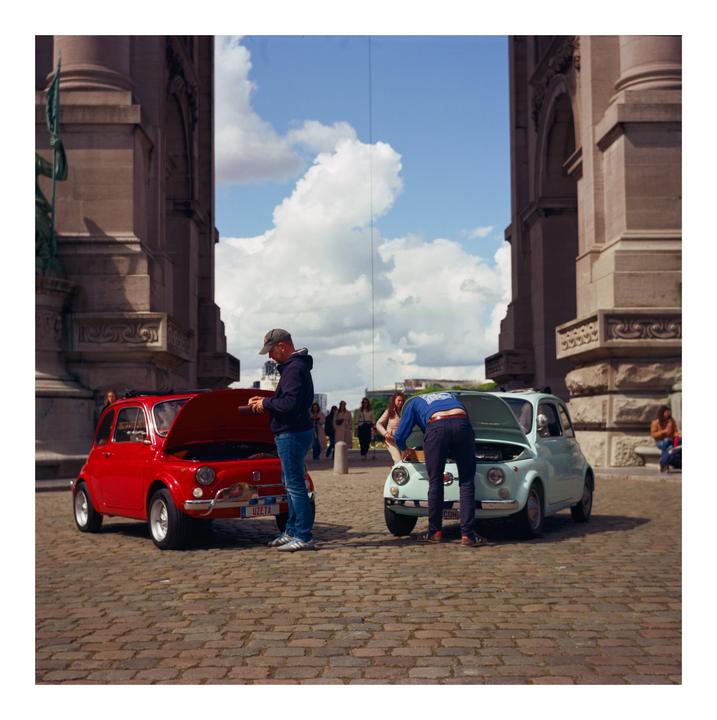 Two men are working on small cars with their hoods open on a cobblestone street next to a large stone building.