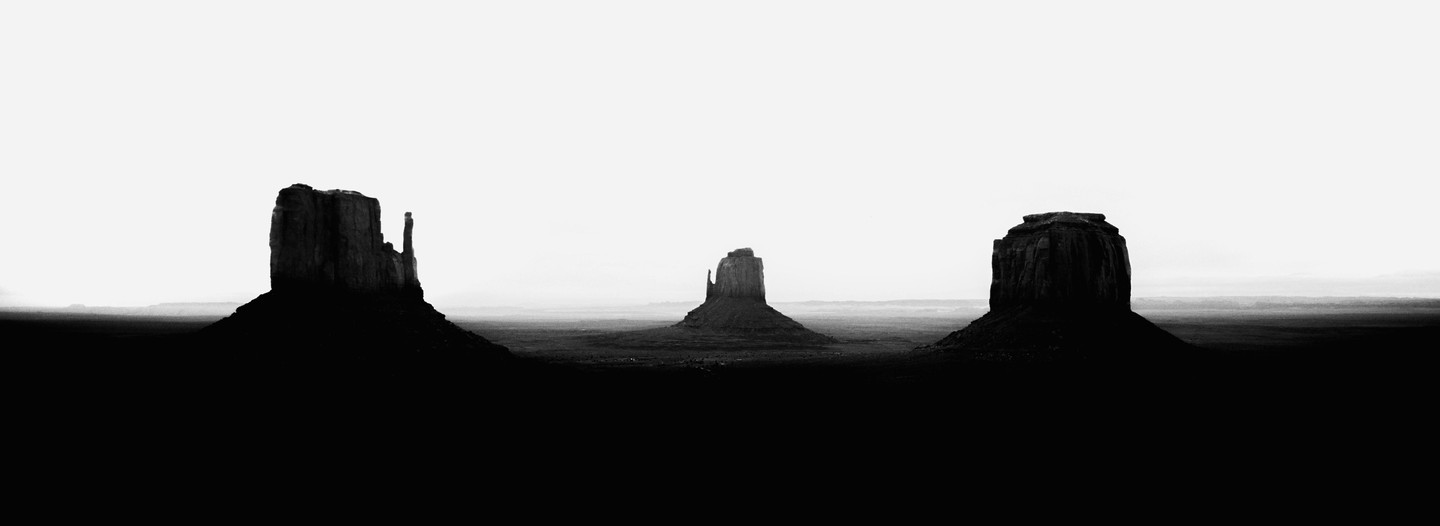 Three large rock formations stand silhouetted against a bright sky in a vast, flat landscape.