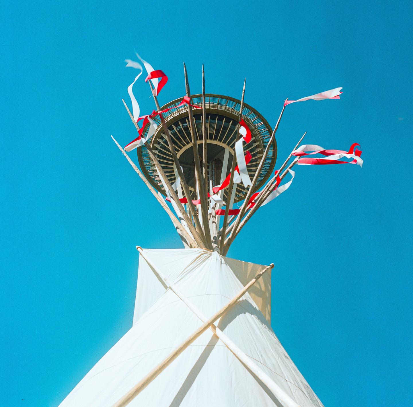 Red and white ribbons adorn a structure resembling a teepee, with the Space Needle visible in the background against a clear blue sky.