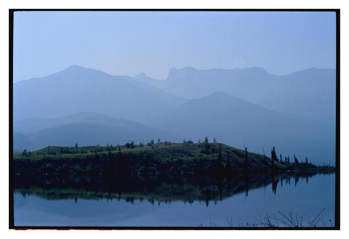A hazy blue landscape shows a lake reflecting a tree-covered hill, with layers of mountains in the background.