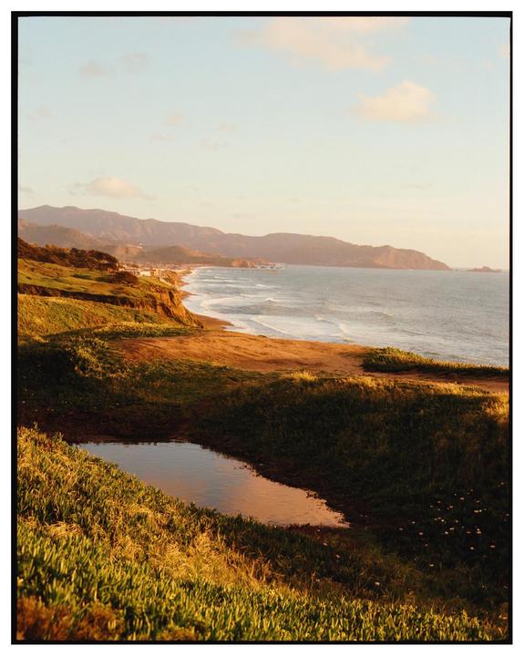 A small pond reflects the sky on a grassy cliff overlooking a beach with waves crashing on the shore and mountains in the distance.