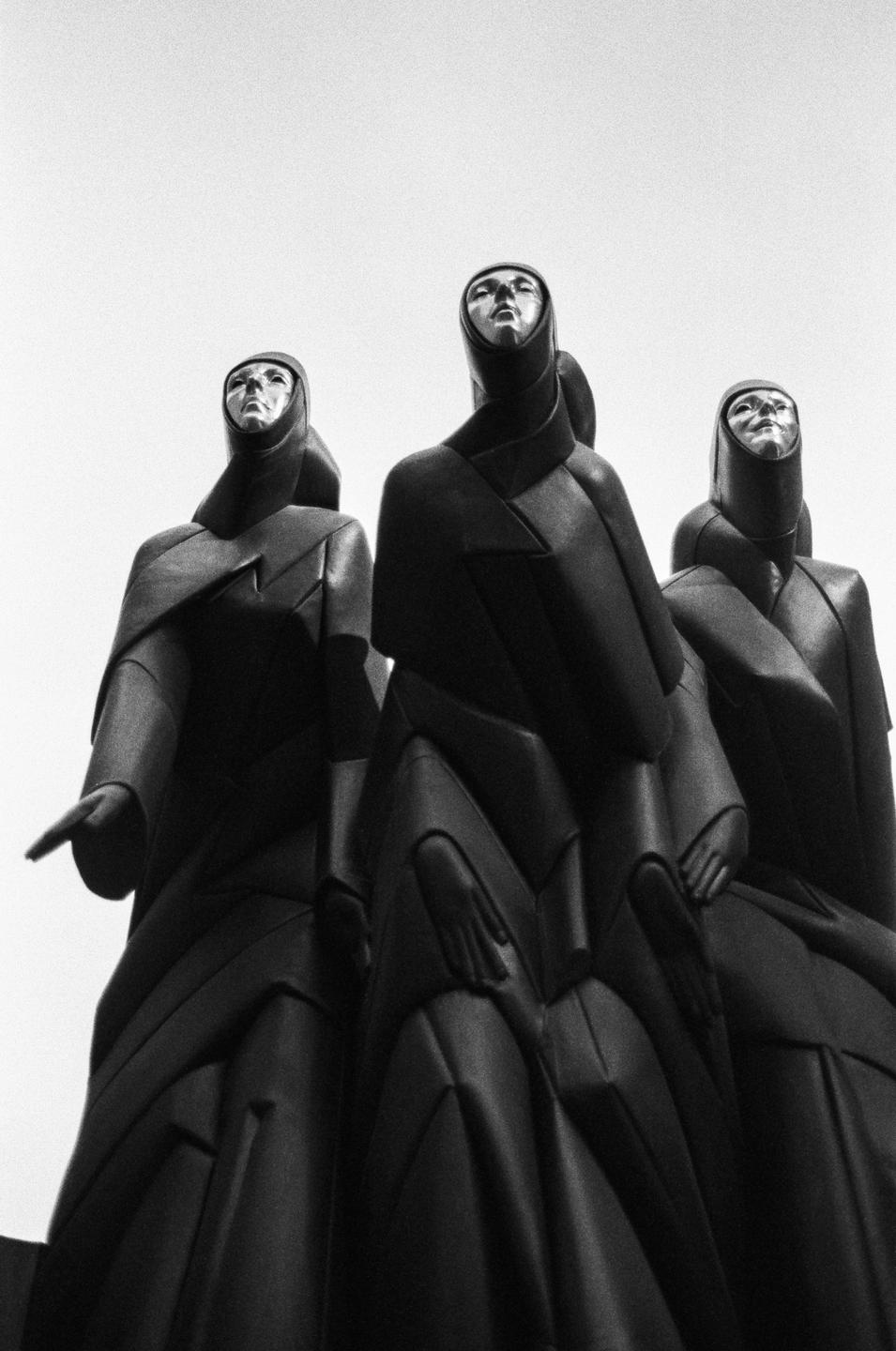 Three statues with metallic faces and dark, flowing robes stand against a bright sky.