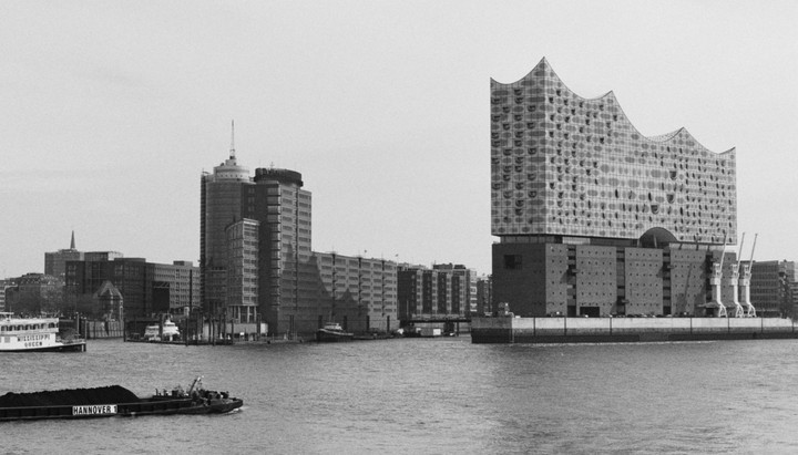 A large, modern building with a wavy roof stands next to a body of water with boats and other buildings in the background.