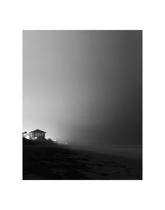 A house with illuminated windows stands on a dark beach at night, with a hazy sky above.