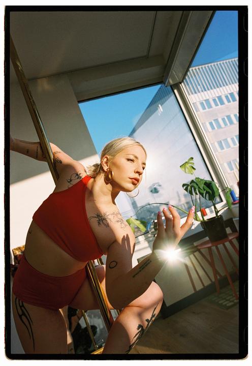 A woman with tattoos and a red outfit poses with a pole in a sunlit room with large windows.