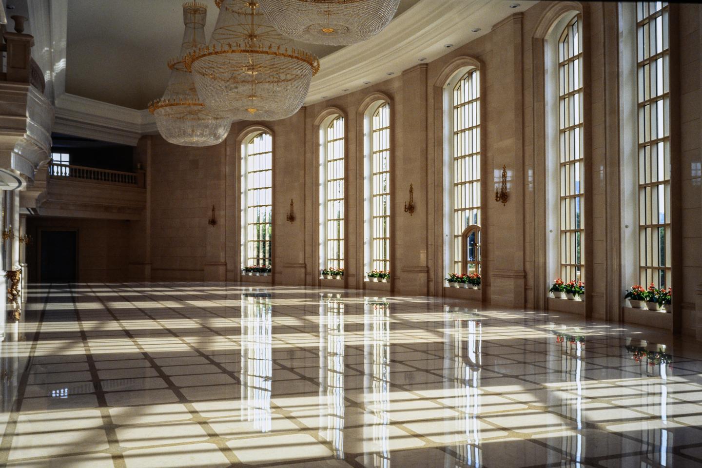 Sunlight streams through tall arched windows, casting long shadows and reflections on a polished marble floor in a grand hall.