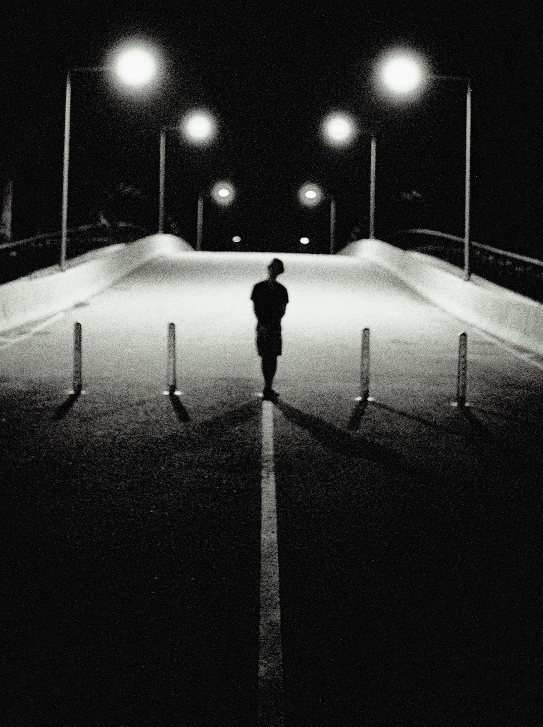 A person stands on a road at night, illuminated by streetlights, with a white line running down the center of the road.