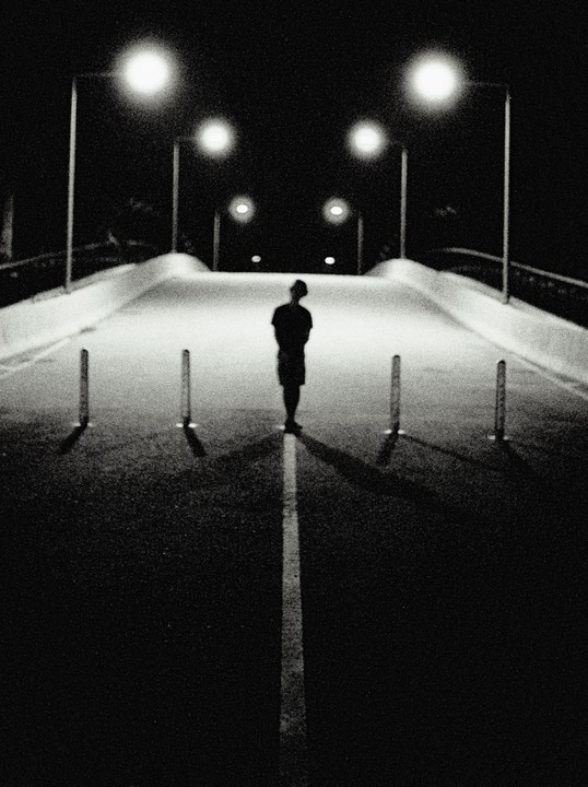 A person stands on a road at night, illuminated by streetlights, with a white line running down the center of the road.