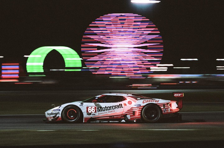 A white race car with red accents speeds past a green arch and a large circular light display at night.