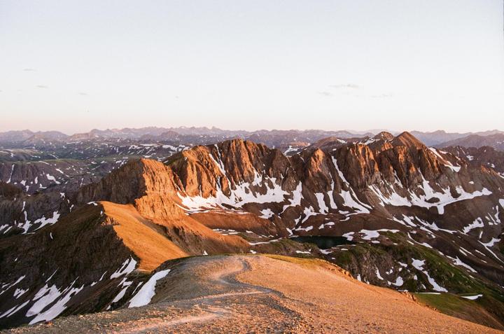 A winding trail leads up a mountain ridge with snow-capped peaks and a small lake visible in the distance under a clear sky.