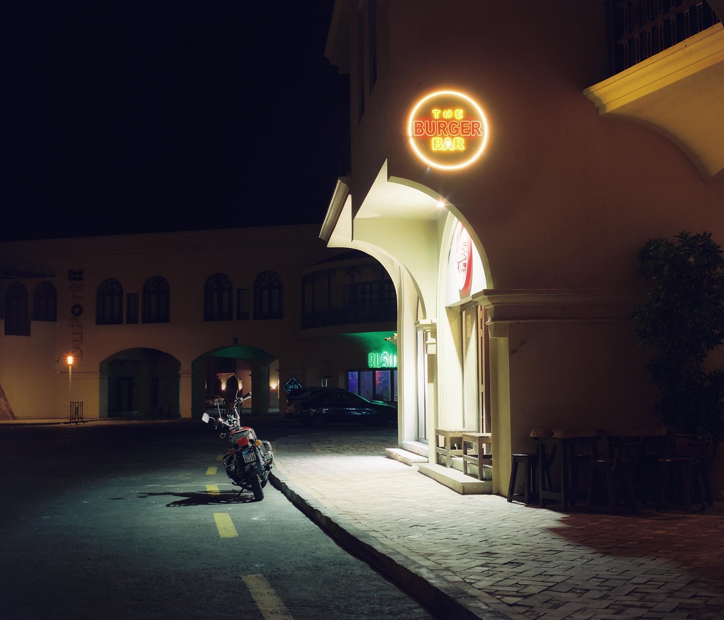 A motorcycle is parked on a street next to a building with a neon sign for "The Burger Bar" at night.