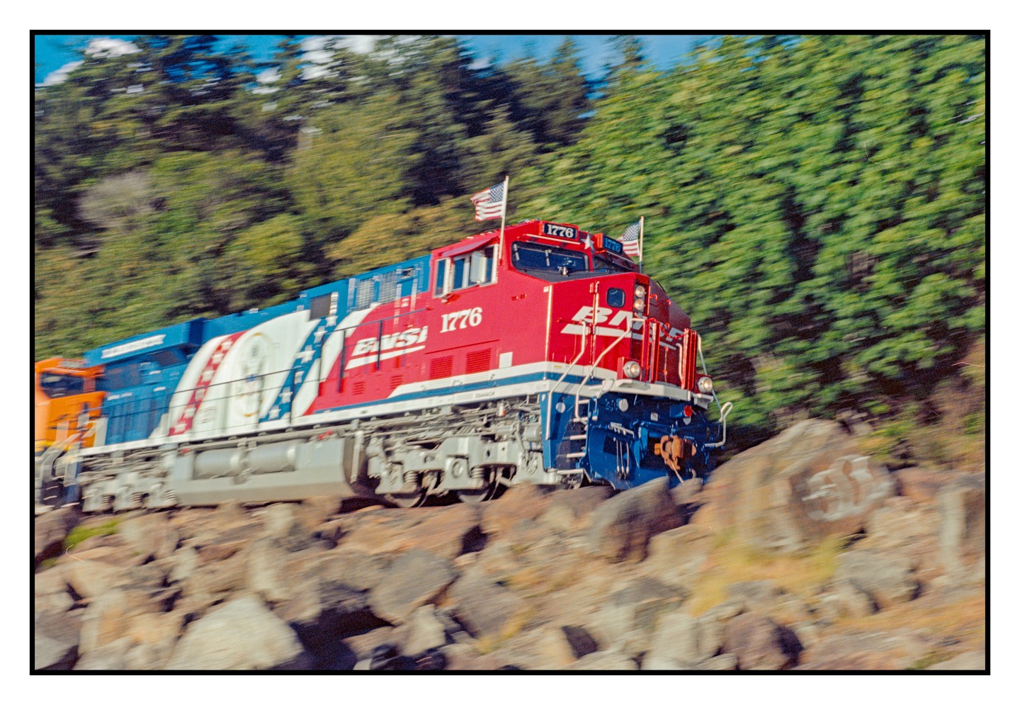 A red, white, and blue train with American flags on top moves past a rocky, tree-lined embankment.