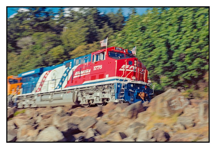 A red, white, and blue train with American flags on top moves past a rocky, tree-lined embankment.