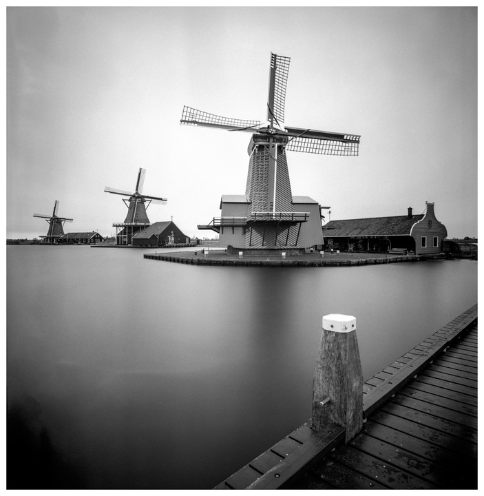 Three windmills stand on the far bank of a body of water, with a wooden pier extending into the foreground on the right.