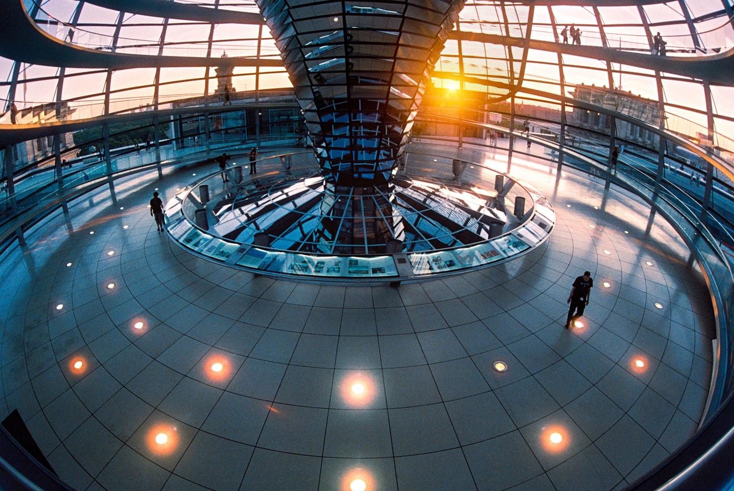 A man walks on a tiled floor inside a modern glass dome with a spiral structure in the center during golden hour.
