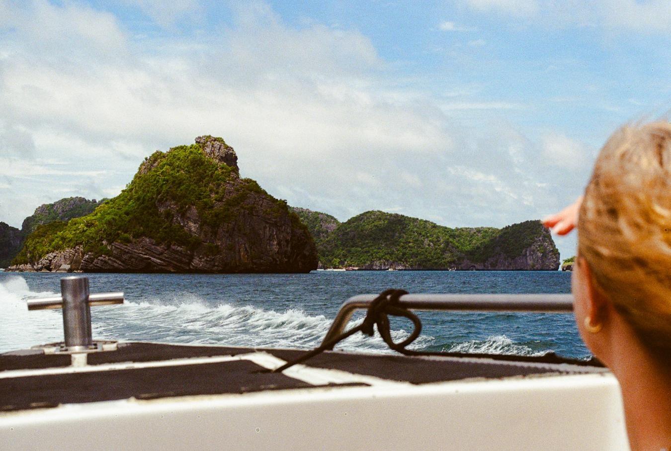 A person on a boat looks out at a tropical island with lush green vegetation and rocky cliffs.