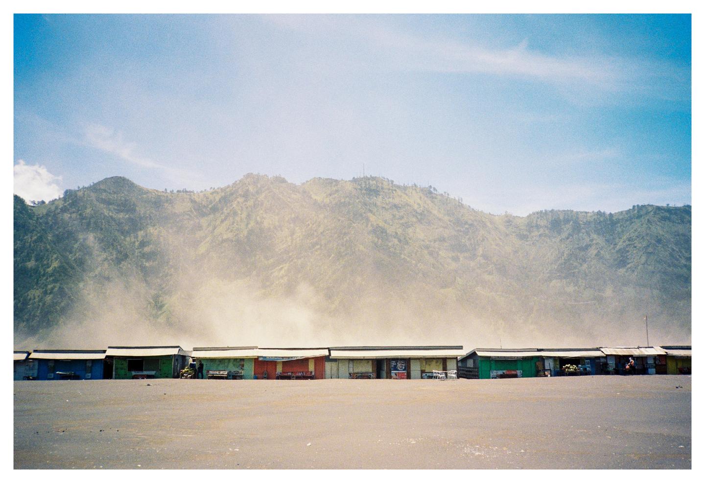 A row of small, colorful shacks sits on a sandy plain with a large, hazy mountain in the background.