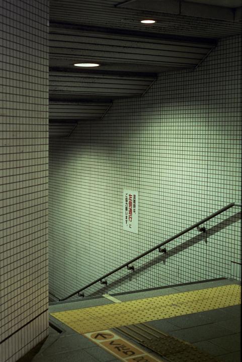 A tiled underground walkway with a handrail and a yellow tactile paving strip on the floor leads down a set of stairs.