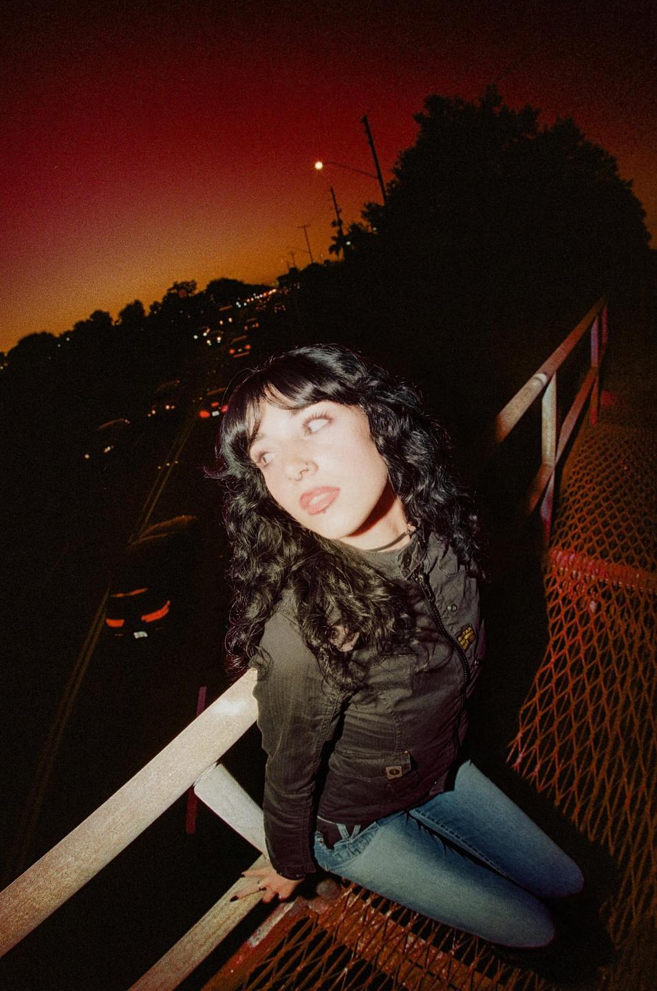 A woman with dark, curly hair and light eyes sits on a metal grate next to a road at night.
