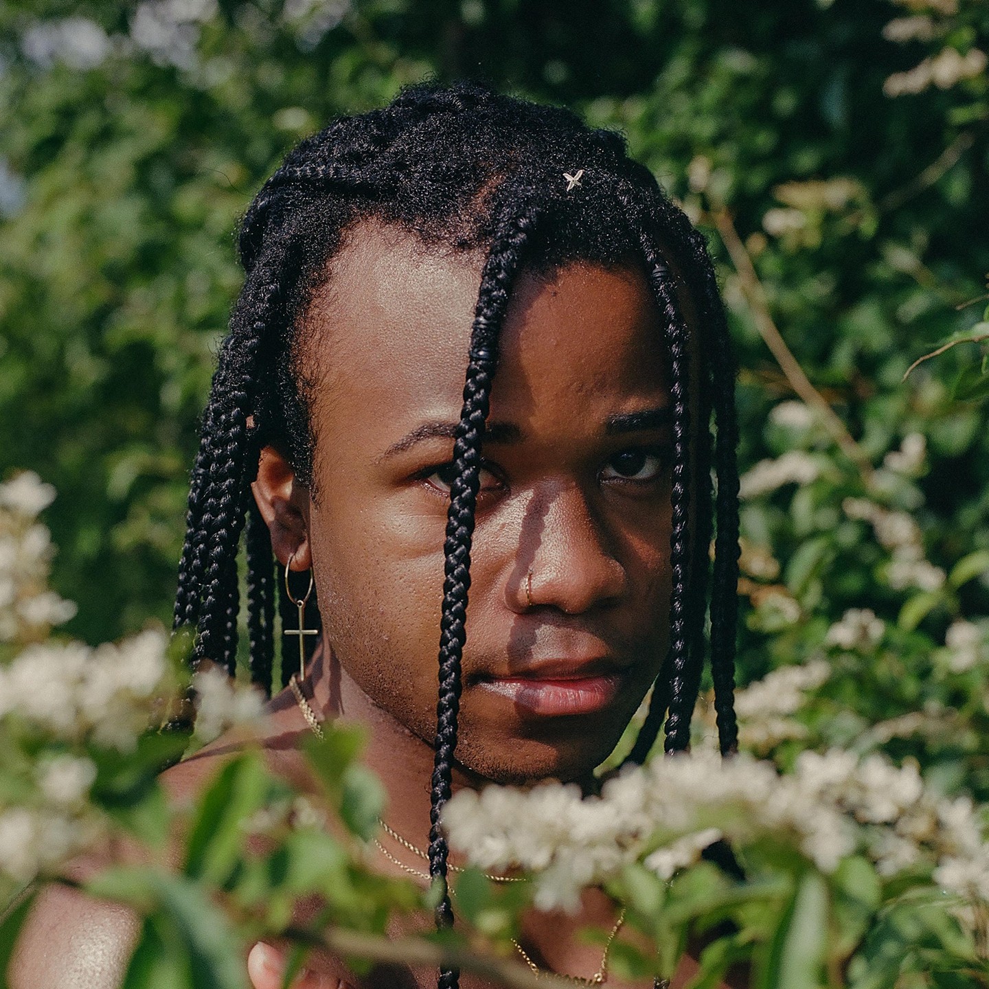 A man with braids and a nose ring looks directly at the viewer from behind white blossoms and green foliage.