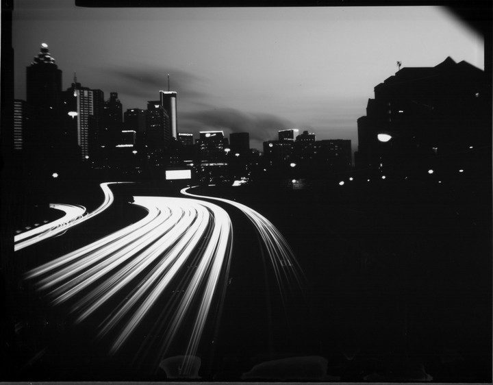 Light trails from vehicles on a highway curve towards a city skyline at dusk.