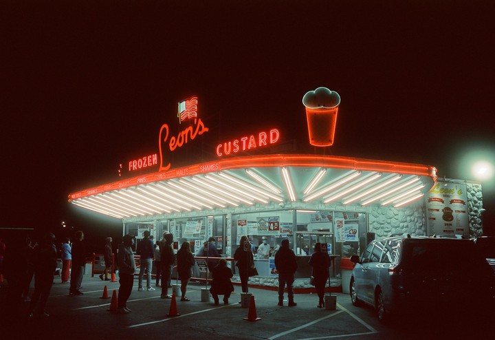A crowd of people stands in line at night outside Leon's Frozen Custard, a brightly lit building with neon signs.