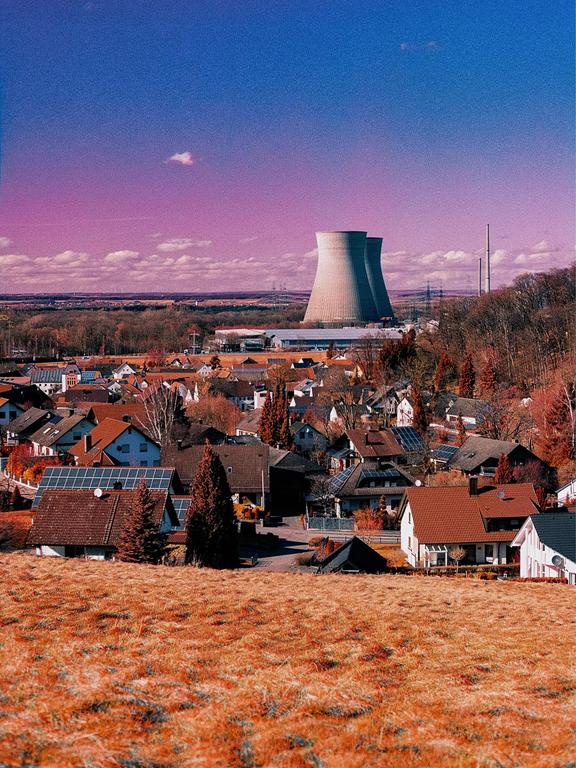 A village with red-roofed houses is nestled in a valley with a power plant in the background under a vibrant blue and purple sky.