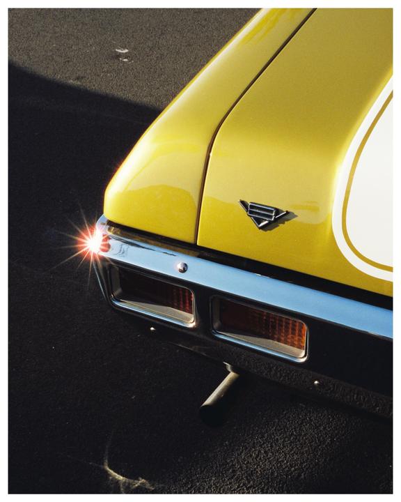 The rear of a yellow car with a white stripe is shown with a bright starburst reflection on the bumper.