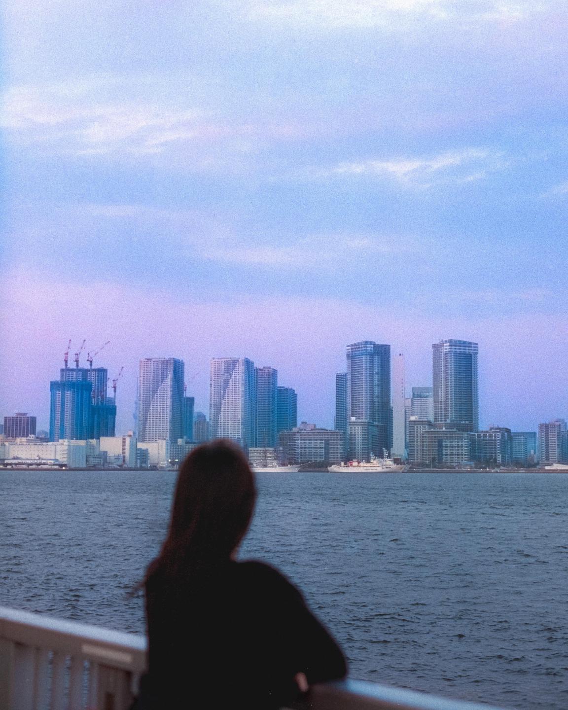 A woman looks out over a body of water at a city skyline under a purple and blue sky.