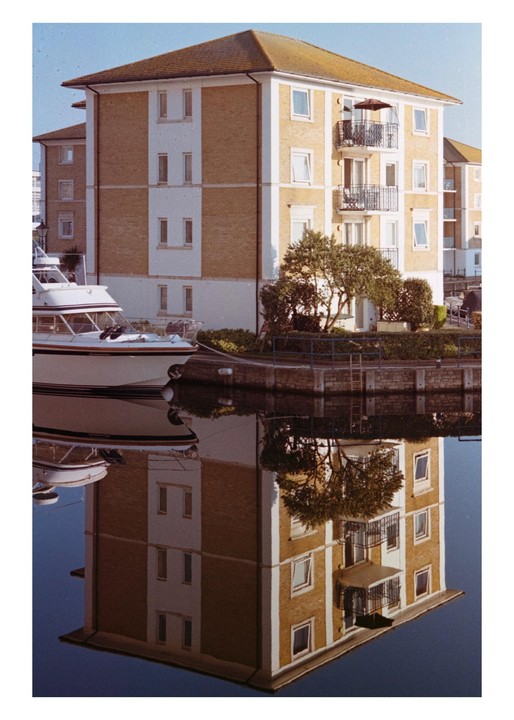 A building with balconies and a boat are reflected in the water of a marina.