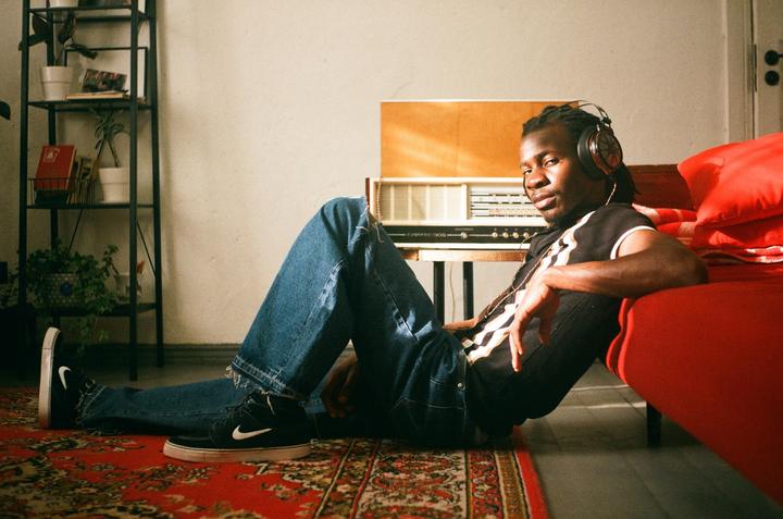 A man wearing headphones and jeans sits on a patterned rug, leaning against a red couch with a vintage radio in the background.