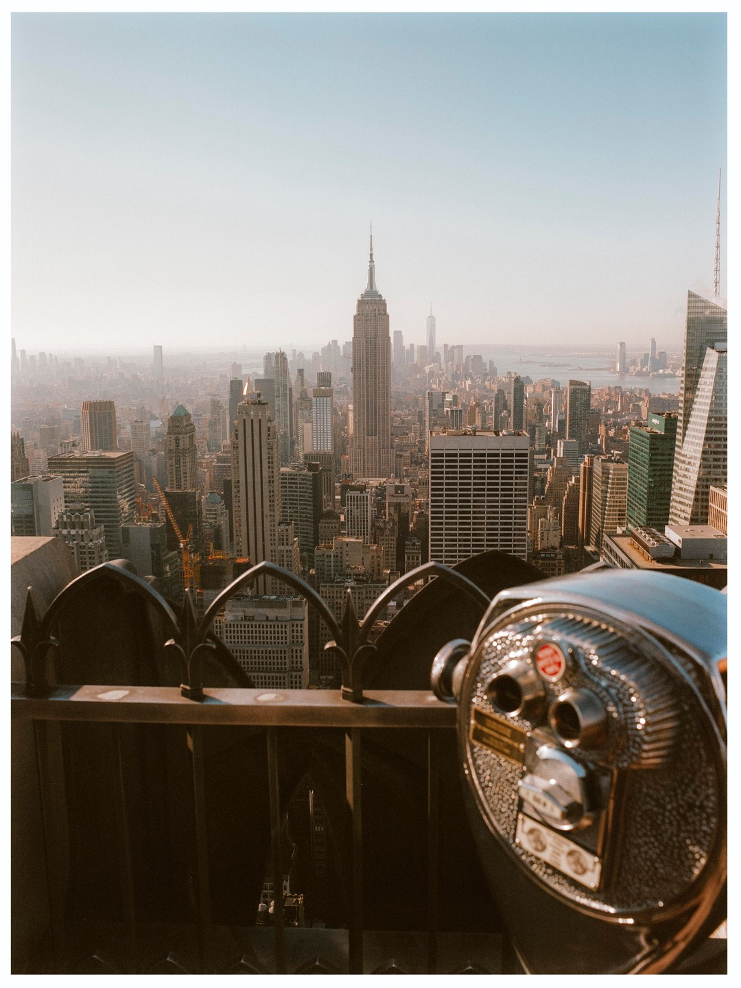 A cityscape of New York with the Empire State Building in the distance is seen from a high vantage point with a coin-operated binocular viewer in the foreground.