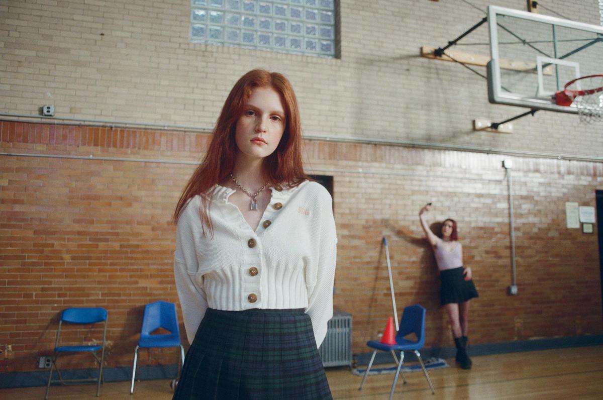 A woman with long red hair stands in a gymnasium while another woman takes a selfie in the background.