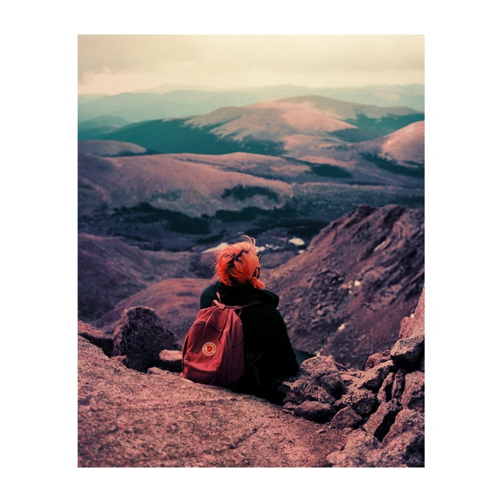 A person with orange hair and a red backpack sits on a rocky mountain overlooking a vast mountainous landscape.