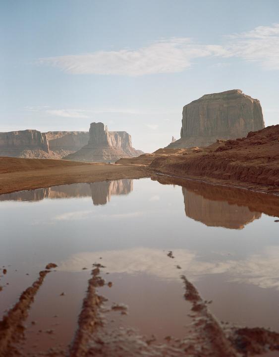 A wide shot shows Monument Valley with a large puddle in the foreground reflecting the sky and the buttes.
