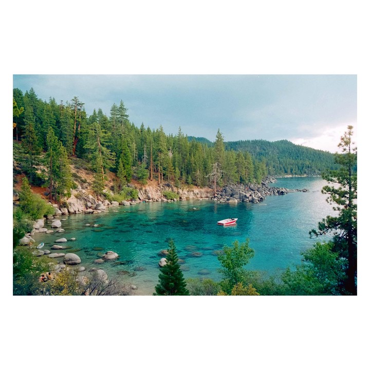 A small red boat floats on clear turquoise water surrounded by a rocky shoreline and dense pine forest under a cloudy sky.