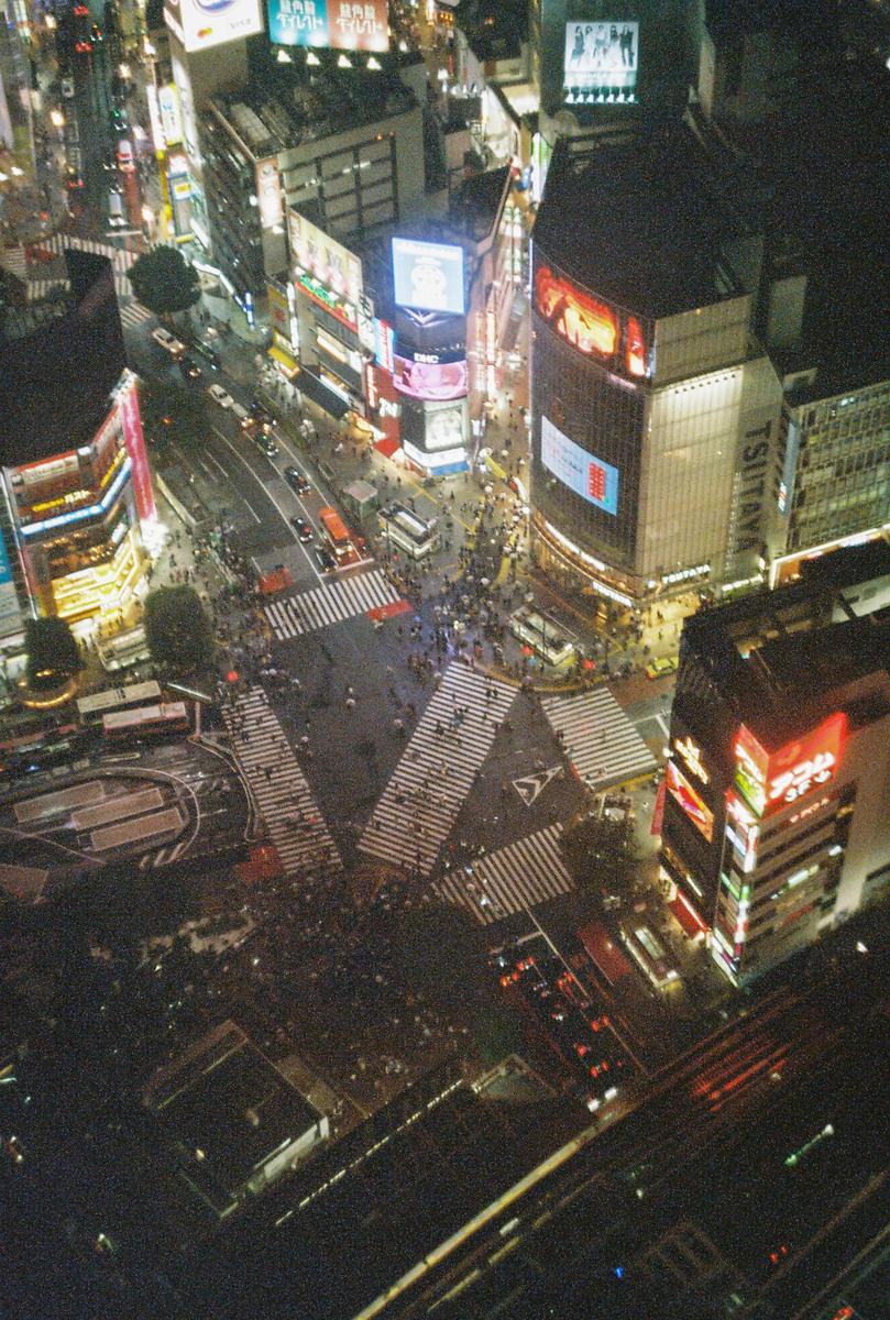 A high-angle view shows the Shibuya crossing at night, with many pedestrians and cars, surrounded by illuminated buildings.