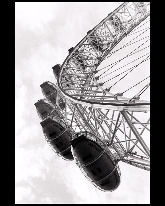 A low-angle black and white shot captures the intricate structure of a ferris wheel against a cloudy sky.