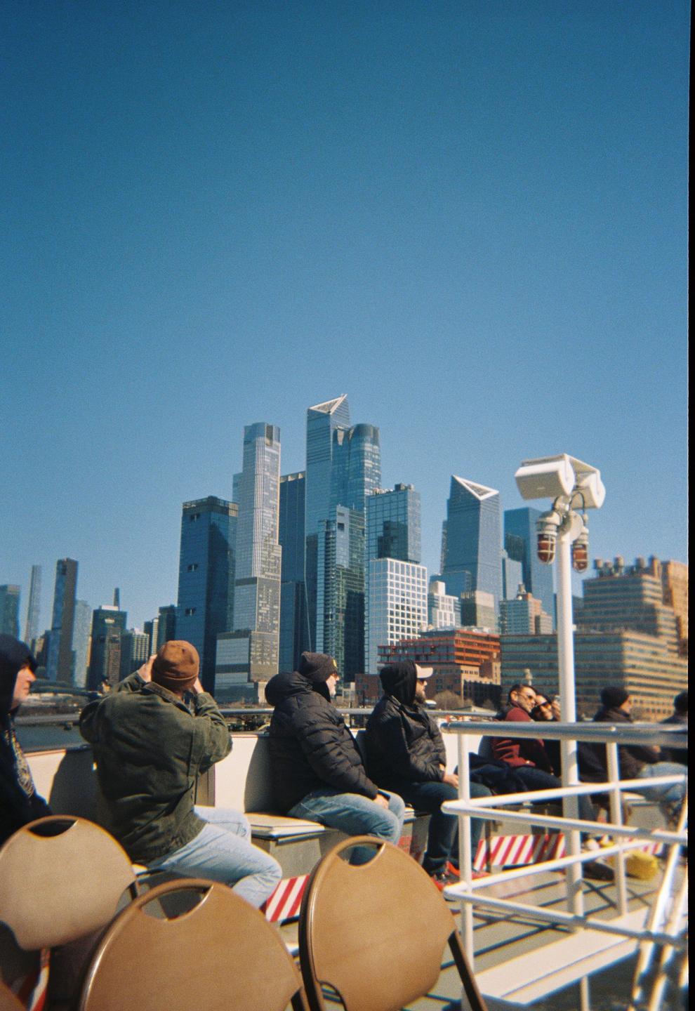 People sit on a boat looking at a city skyline with tall buildings under a clear blue sky.