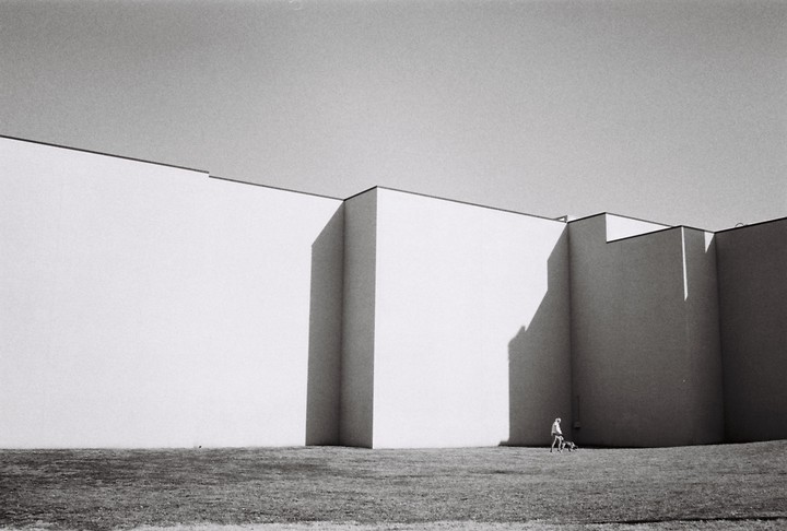 A woman walks her dog on a grassy area next to a large, modern building with strong shadows.