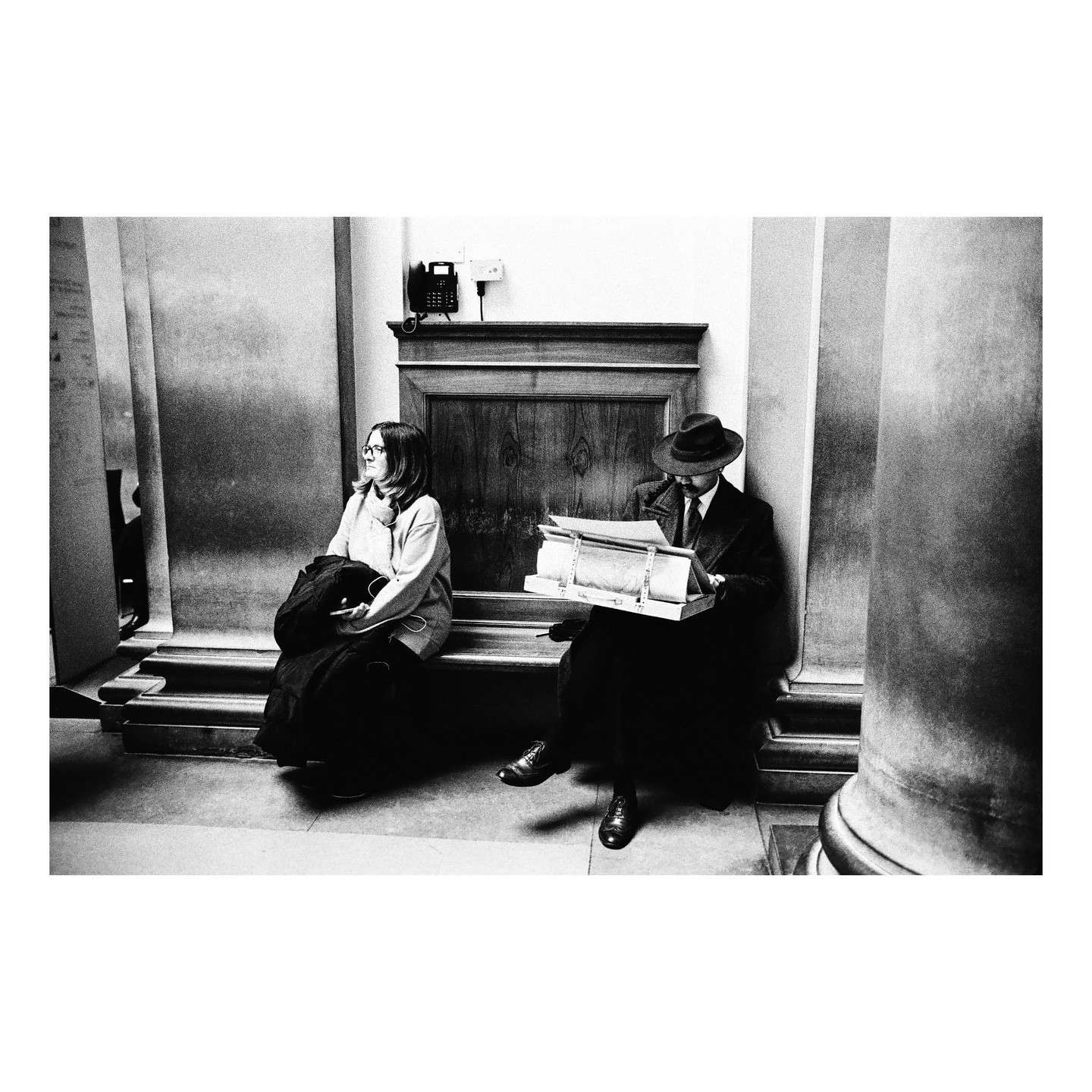 A woman and a man in a hat sit on a bench, with the man looking at papers in a box.
