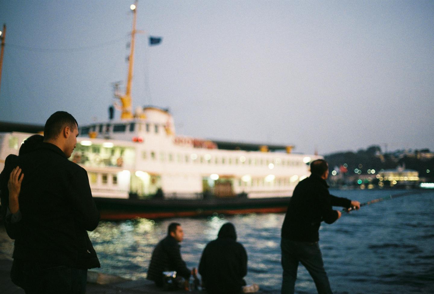 A group of men are gathered by the water with a large ferry boat in the background.