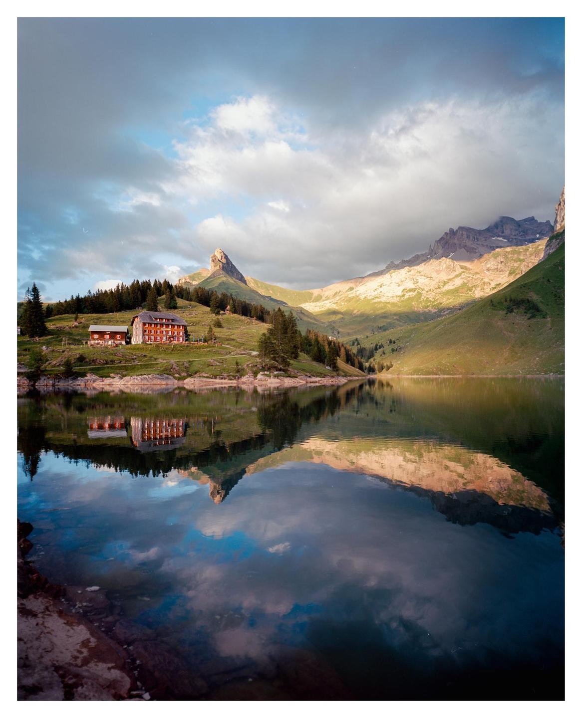 A red building sits on a grassy hill next to a lake, with mountains and clouds reflected in the water.